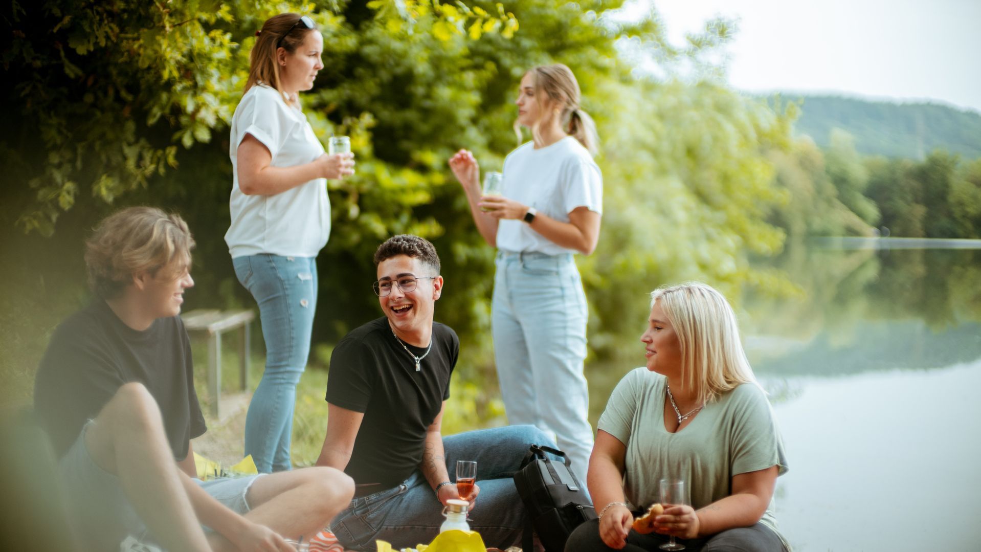 Das Bild zeigt mehrere Menschen bei einem Picknick. Im Hintergrund ist ein See zu sehen. Zwei Menschen stehen, drei sitzen auf einer Decke auf dem Boden und unterhalten sich. 