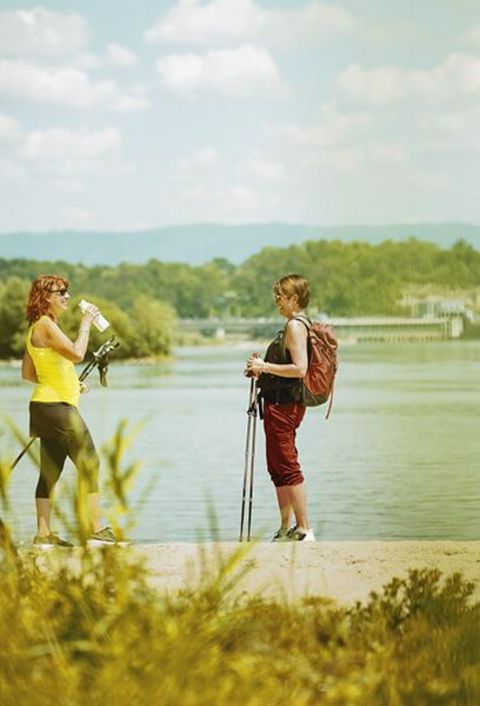 Zwei Damen mit Wanderstöcken und Freizeitkleidung machen eine Pause vor dem Rhein, im Hintergrund das Wasserkraftwerk von Rheinfelden.